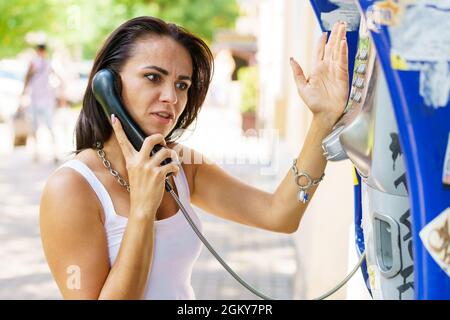 Positive brunette femme tenant des appels de récepteur de téléphone par téléphone de papeterie dans la rue, touriste parlant sur le téléphone de paye travaillant avec des pièces, heureux avec la communication internationale le jour ensoleillé d'été Banque D'Images