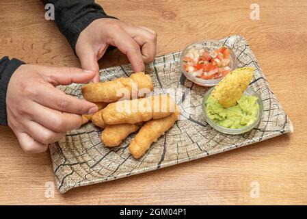 Les mains du chef plaçant une portion de tequeños vénézuélien frits sur une plaque en rondins accompagnée d'un petit guacamole avec des chips tortilla et pico de gallos Banque D'Images