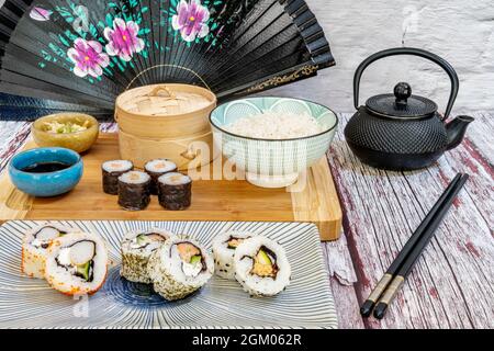 Gros plan de l'uramaki, du rouleau de californie, du saumon maki et du bol à riz avec théière noire, baguettes noires et éventail sur table rouge et blanche Banque D'Images