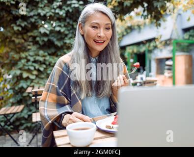 Bonne dame asiatique mange des toasts avec des fraises en regardant la vidéo par ordinateur portable à la table à l'extérieur Banque D'Images