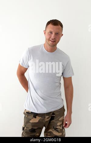 Positif jeune homme en kaki militaire pantalon main derrière et t-shirt gris debout sur fond blanc et regardant l'appareil photo Banque D'Images