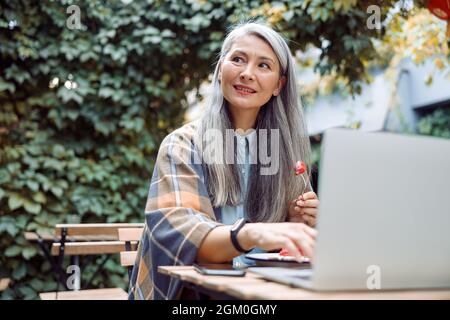 Une femme asiatique d'âge moyen attentionnés avec un dessert se trouve devant un ordinateur portable sur une table à l'extérieur Banque D'Images