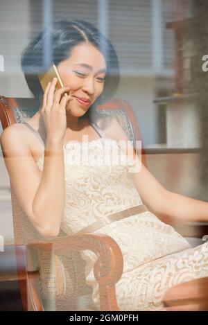 Souriante belle jeune femme asiatique appréciant parler au téléphone avec un ami ou un membre de la famille lors de passer un week-end à la maison Banque D'Images