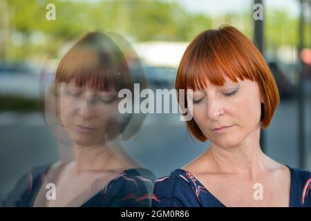 Femme abattue avec une expression triste et les yeux bagués debout contre une fenêtre de magasin dans la ville reflétée dans le verre dans une tête de gros plan Banque D'Images