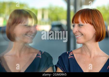 Une femme joyeuse et amusante qui regarde son propre reflet dans une grande fenêtre en verre de plaque commerciale dans une rue calme de la ville avec un sourire heureux dans un gros plan Banque D'Images