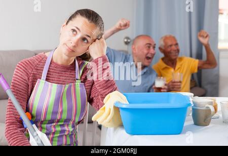 Femme fatiguée pendant les travaux ménagers, homme âgé avec homme regardant la télévision Banque D'Images