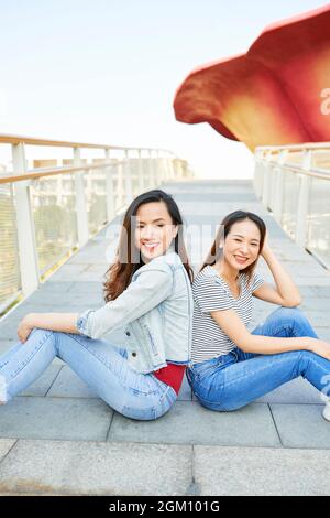 De belles jeunes amies vietnamiennes assises dos à dos sur le pont et souriant à la caméra Banque D'Images