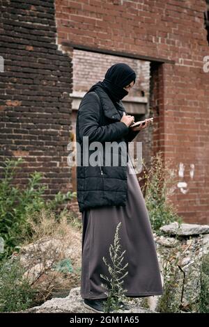 Réfugiée du Moyen-Orient en hijab et longue tenue debout contre un bâtiment abandonné et utilisant le téléphone Banque D'Images