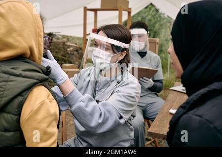 Jeune femme médecin dans un écran facial de protection vérifiant la gorge de l'enfant réfugié à l'extérieur Banque D'Images