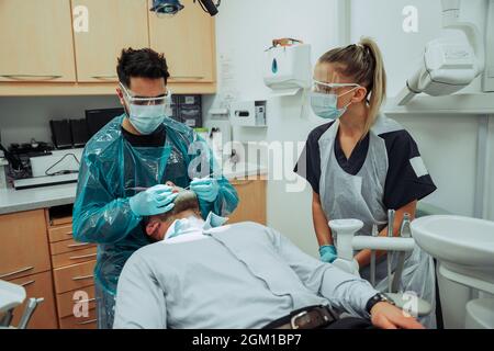 Dentistes blancs, hommes et femmes, debout dans la salle des médecins tout en agissant sur les dents des clients mâles Banque D'Images