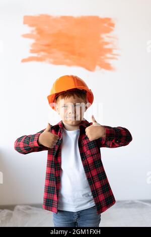 Un enfant de fils se tient dans un casque orange près d'un mur blanc et a levé ses pouces vers le haut pointant vers un endroit pour le texte. Réparation à domicile par vous-même. Banque D'Images