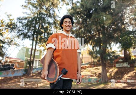 Skateboarder insouciant tenant son skateboard dans un parc urbain. Adolescent joyeux souriant à la caméra tout en se tenant dehors. adolescent sportif wea Banque D'Images