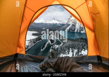 Homme de voyage se reposant et profitant de la vue sur la montagne enneigée et le lac à l'intérieur d'une tente orange en hiver au parc national Yoho, Canada Banque D'Images