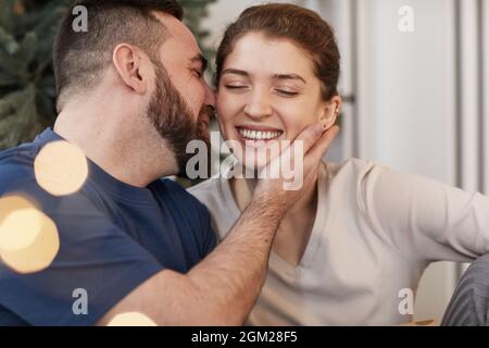Joyeux jeune homme barbu embrassant la femme bien-aimée avec les yeux fermés sur le nouvel an, effet de lumière de lentille Banque D'Images