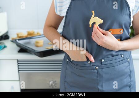 Femme avec un tablier et une main dans la poche tenant un cookie en forme de chat d'halloween. Cuisine domestique,. Copier l'espace. Banque D'Images