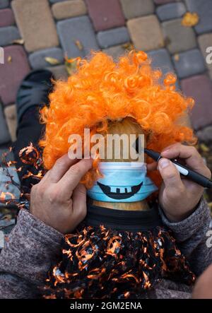 Les mains mâles attirent les yeux avec un marqueur noir sur une citrouille dans un masque médical bleu. Se préparer à des vacances terribles. Fêtez Halloween pendant la COVID-1 Banque D'Images