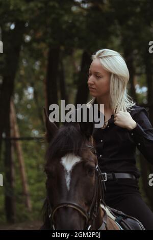 Une fille blonde en profil à cheval noir contre une forêt verte. Une fille est à cheval brun un jour d'automne. Banque D'Images