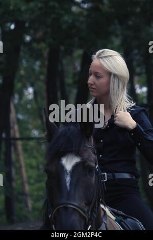 Une fille blonde en profil à cheval noir contre une forêt verte. Une fille est à cheval brun un jour d'automne. Banque D'Images