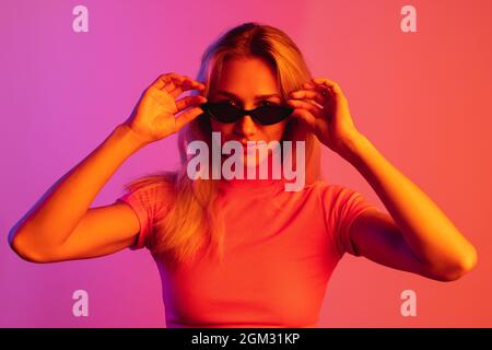 Image courte d'une jeune fille élégante portant des lunettes de soleil tendance isolées sur fond dégradé dans des lumières fluo. Clubbing Banque D'Images