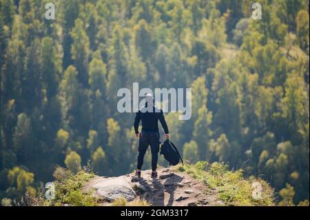 sportif actif et en bonne santé, homme en tenue de sport noire et casquette tenant un sac à dos debout en haute montagne avec des arbres de forêt en arrière-plan Banque D'Images