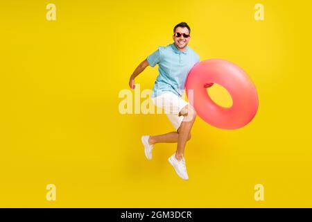 Photo de joli doux jeune gars habillé bleu t-shirt sombre oculaire courant jumping tenant anneau gonflable isolé couleur jaune fond Banque D'Images