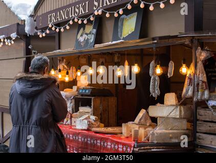 Un homme se tenant en face du marché de vacances de rue décoré de lumières jaunes vendant du fromage et des saucisses Banque D'Images