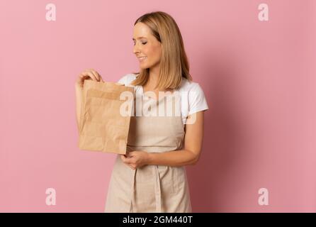 Souriante bonne jeune femme dans un tablier tenant marron vide sac en papier artisanal vierge pour emporter maquette isolée sur fond rose Banque D'Images