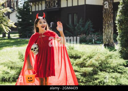 fille émerveillée en diable halloween costume de la main en se tenant avec le crâne et le seau de bonbons près de la maison floue Banque D'Images