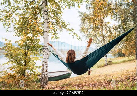 La jeune femme leva les bras avec joie pendant qu'elle balança dans un hamac entre les bouleaux sur la rive du lac de montagne. Loisirs en plein air à l'extérieur de la ville Banque D'Images