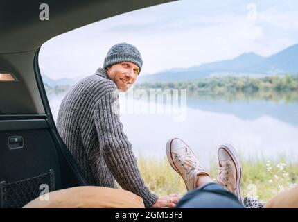 Un homme d'âge moyen vêtu de vêtements tricotés chauds près de la voiture, souriant devant une femme dans un coffre qui jouit d'une vue sur le lac. Confortable début de l'automne Banque D'Images