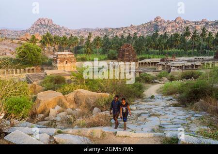 Hampi, Karnataka, Inde: Un homme et une femme marchent au coucher du soleil parmi les anciennes ruines de Vijayanagar. Temple Achyutaraya du XVIe siècle en arrière-plan. Banque D'Images