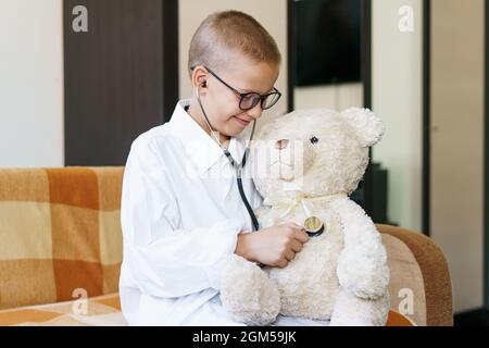 Mignon petit garçon caucasien jouant un jeu drôle comme le docteur tenant stéthoscope à l'écoute de préchooler jouet prétendant être infirmière traiter patient moelleux à la maison. Dans une robe de chambre et un masque médical. Banque D'Images