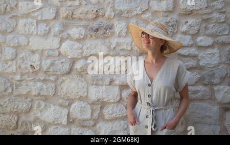 Portrait d'une belle blonde gaie portant une robe d'été et un chapeau d'été, debout devant un vieux mur médiéval en pierre. Été Banque D'Images