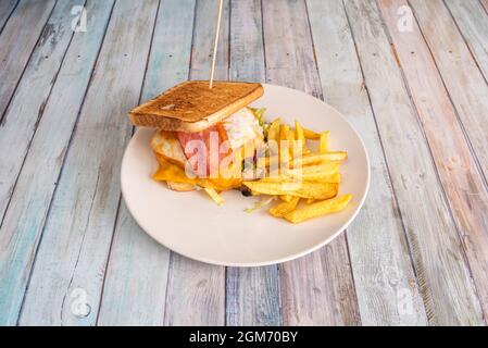 Sandwich maison avec œuf frit, bacon et tranches de cheddar, laitue et viande de poulet, garniture de pommes de terre frites maison sur plaque blanche et Banque D'Images