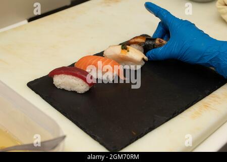 Plateau à sushis nigiri avec des morceaux de thon rouge, de saumon norvégien, de babeurre et d'anguille fumée placés par la main gantée du chef en plastique bleu Banque D'Images