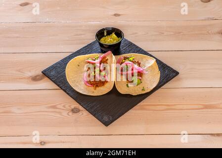 Deux tacos mexicains de cochinita pibil avec un bol de guacamole sur une plaque d'ardoise noire sur une table en bois Banque D'Images