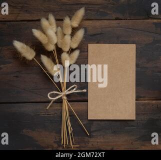 Carte en papier kraft vierge sur table en bois brun avec bouquet de fleurs séchées de côté, vue du dessus. Maquette de carte d'invitation de boho Banque D'Images