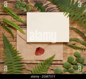 Carte en papier vierge avec enveloppe scellée et joint en cire rouge posé sur une table en bois marron avec des feuilles de fougères autour de la vue de dessus. Maquette tropicale avec invi Banque D'Images