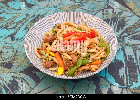 Nouilles udon de chubby sautées de poivrons rouges et verts, viande de porc, fleurs comestibles sur un bol râpé Banque D'Images