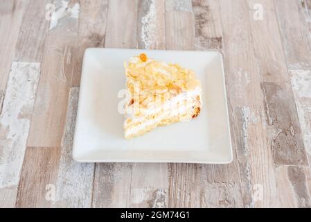 Portion de gâteau aux amandes et crème avec gâteau éponge et deux couches de crème blanche sur plaque carrée et table en bois Banque D'Images