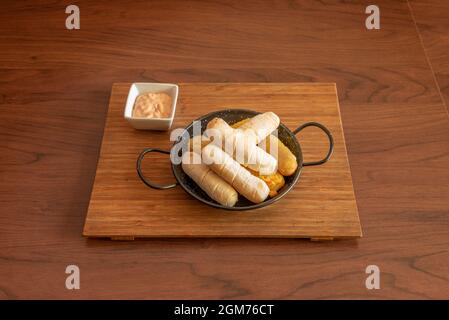 Service de tequeños de différentes saveurs dans une petite poêle noire et sauce à tremper sur un panneau de bambou Banque D'Images