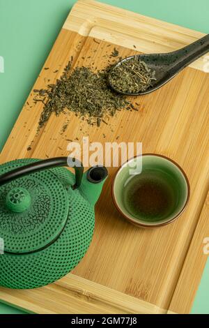 Vue de dessus image d'un ensemble traditionnel de thé vert et d'une cuillère en porcelaine noire avec du thé sec sur une table en bambou brun et un fond vert Banque D'Images