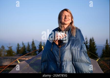 Une femme voyageur enveloppée dans un sac de couchage pour une tasse de café et souriant, debout près d'une tente de camp avec un ciel bleu sur le fond. Concept de randonnée, de voyage et de camping. Banque D'Images
