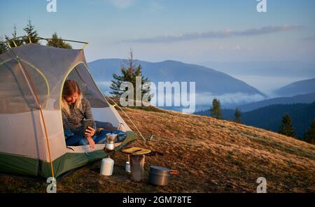 Femme voyageur assis dans une tente de camp et prenant une photo du brûleur à gaz touristique et de la bouilloire. Randonneur féminin utilisant un téléphone portable tout en se reposant dans une tente touristique sur une colline herbeuse avec montagne et ciel sur fond. Banque D'Images