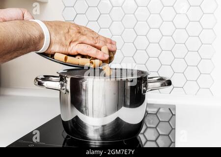 Les mains des adultes versent des pâtes italiennes dans une casserole d'eau bouillante sur une table de cuisson à induction Banque D'Images