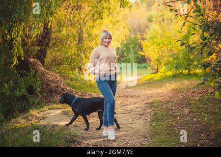 Une femme blonde avec une coupe courte traverse la forêt dans un pull tricoté et un grand chien noir rottweiler. Randonnée en plein air dans la forêt d'automne Banque D'Images