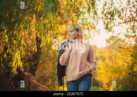 Une femme blonde avec une coupe courte traverse la forêt dans un pull tricoté et une veste en cuir sur son épaule. Randonnée en plein air dans les forts d'automne Banque D'Images