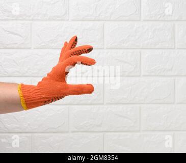 main femelle dans un gant de protection orange de travail contre le fond d'un mur de brique blanche, la main tient conditionnellement un objet Banque D'Images