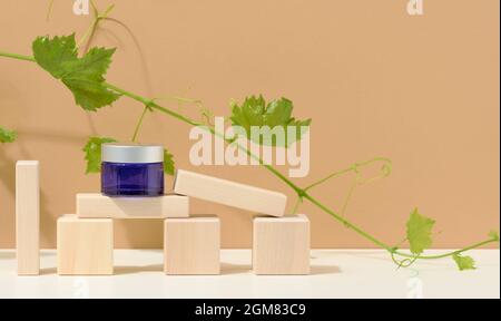 produits cosmétiques dans un pot en verre bleu avec un couvercle gris sur un podium en bois en cubes, derrière une branche de raisins avec des feuilles vertes. Vide pour le branding Banque D'Images