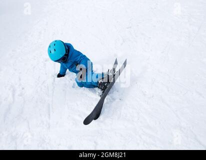 un enfant dans un casque, des lunettes, des skis et une combinaison bleue se trouve dans la neige, est tombé. Cours de ski pour enfants à l'école de ski. Animations hivernales pour Banque D'Images